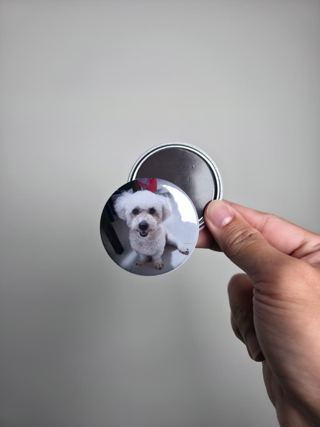 Hand holding a round photo magnet with a dog's face printed on it against a gray background