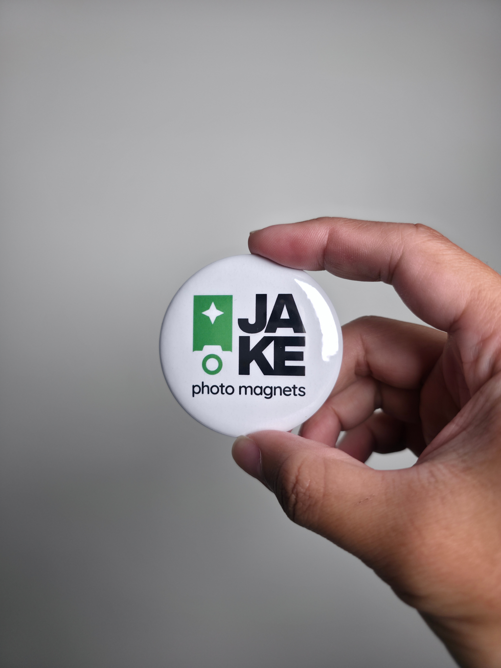 Hand holding a round white magnet with 'JAKE photo magnets' logo against a gray background
