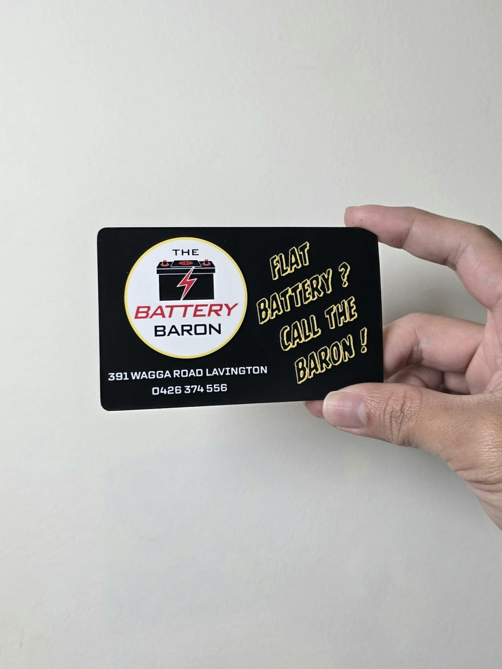 Hand holding a card with 'The Battery Baron' branding and contact information.