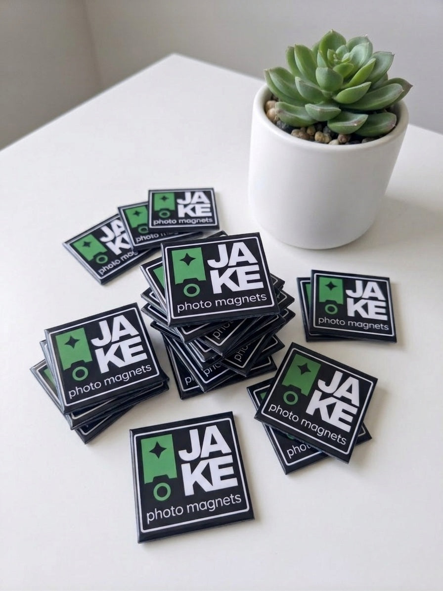 Stack of JAKE photo magnet stickers on a white surface with a potted plant in the background.