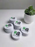 Stacks of white photo magnets with 'JAKE' branding on a light surface with a plant in the background.