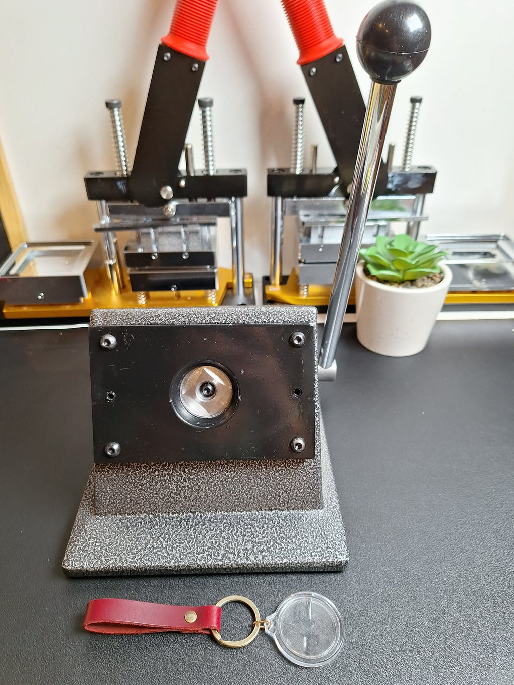 Press machine with red handles on a gray surface, with a keychain and clear plastic item nearby.
