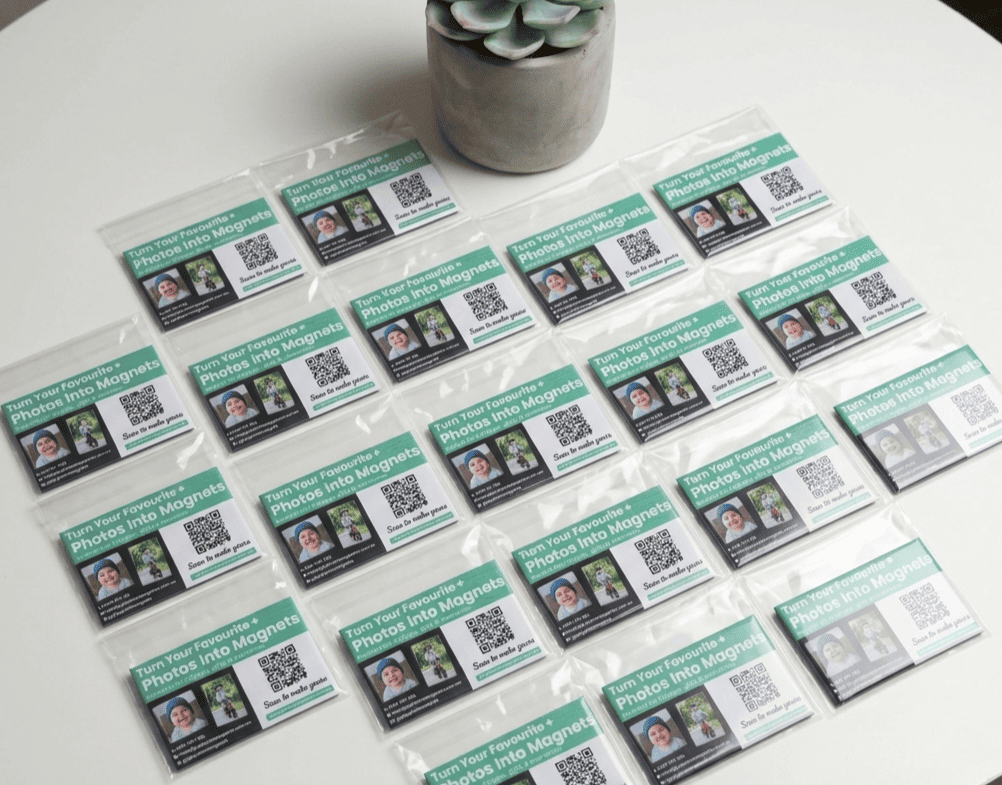 Multiple photo magnets with QR codes on a white surface with a plant in the background.